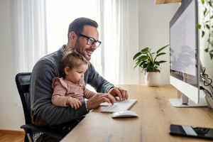 Ein Vater - Unternehmer tippt einen Bericht, während er seine Tochter im Homeoffice auf dem Schoß hält. iStock-1459014790