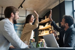 Zwei fröhliche junge Geschäftsleute, die High-Five geben, während ihre Kollegen sie anschauen und lächeln. iStock-1340692189