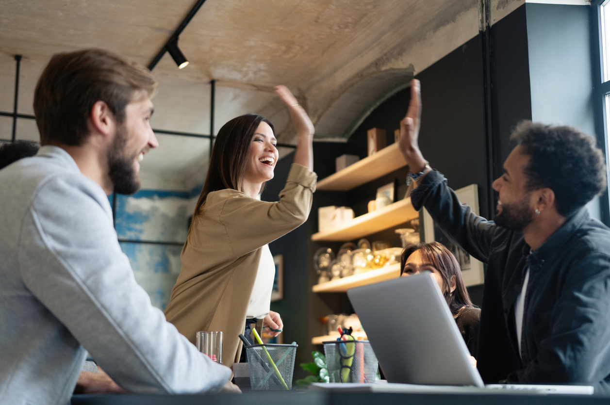 Zwei fröhliche junge Geschäftsleute, die High-Five geben, während ihre Kollegen sie anschauen und lächeln. iStock-1340692189